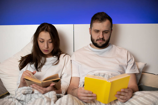 Όμορφο νεαρό ζευγάρι κάθεται στο κρεβάτι διαβάζοντας βιβλία στο σπίτι στην κρεβατοκάμαρα - Φωτογραφία, εικόνα