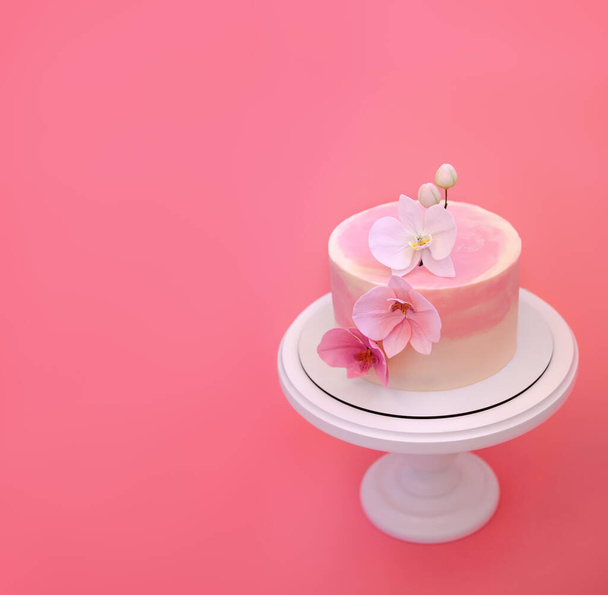 Vaaleanpunainen kakku seisoo pyöreä valkoinen seistä vaaleanpunainen tausta. Kakku koristeltu sokeri kukkia orkideat. Kaunis jälkiruoka koristeltu kukilla. - Valokuva, kuva