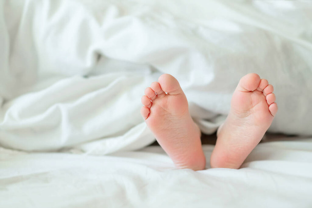 Heels and fingers on the bed. Heels and feet. Children's legs. Barefoot on the bed. Baby legs in bed. Children rest focused on bare feet. Baby bare feet peeking out from under a white blanket. - Photo, image