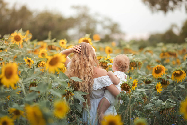 Mladá krásná matka se drží za ruce malé kudrnaté dcerky. Mateřství. kudrnaté maminka a malá dcera na slunečnicovém poli. - Fotografie, Obrázek