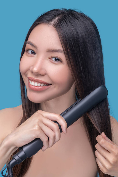 Smiling long-haired woman using a straightening iron - Foto, Imagen