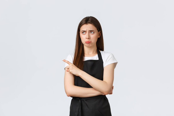 Small business, employees and coffee shop concept. Jealous and upset cute barista in black apron pouting unsatisfied and pointing finger left at sad disappointing news, white background - Photo, Image