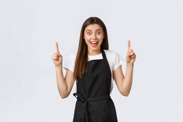 Small business, employees and coffee shop concept. Confient pretty barista in black apron pointing fingers up. Cafe employee showing advertisement. Cheerful saleswoman recommend special promo - Photo, Image