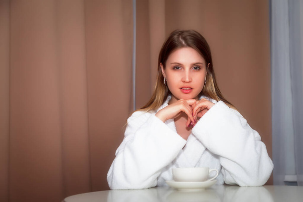 Girl of Slavic appearance in coat with white Cup is sitting at table. Female takes actor's emotions. Young charming woman is sitting in hotel room and drinking coffee or tea. Relax concept - Фото, изображение