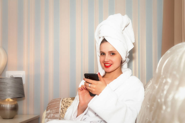Woman with phone on sofa of hotel room after shower. Pretty female of Slavic appearance in dressing gown and towel on her head. Happy lady. Lesson with smartphone. Cute girl after washed - Photo, image