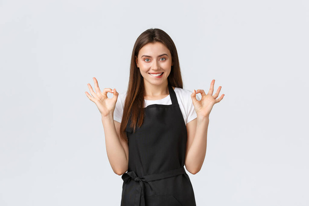 Grocery store employees, small business and coffee shops concept. Satisfied happy female barista in black apron showing okay sign and biting lip tempting. Pleased saleswoman recommend buy online - Foto, imagen