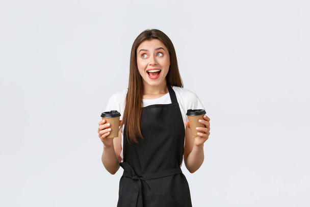 Grocery store employees, small business and coffee shops concept. Friendly cheerful smiling barista holding two cups of drinks, looking left dreamy as serving order to clients, white background - Foto, immagini