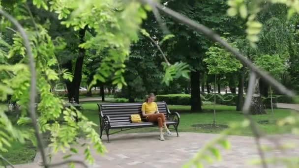 The camera through the branches of trees follows a teenage girl sitting on a bench in a city Park with a phone in her hands - Materiaali, video
