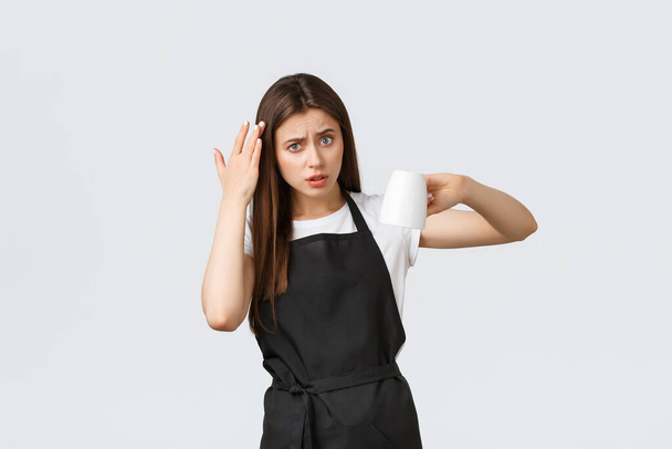 Grocery store employees, small business and coffee shops concept. Confused and displeased woman pointing at head and showing empty cup with disappointed grimace, white background - Photo, Image