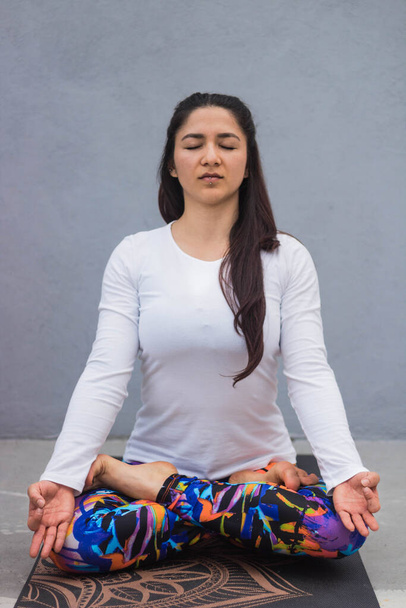 Женщина в белой футболке медитирует, скрестив ноги на коврике для йоги
 - Фото, изображение