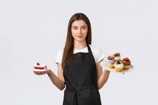 Zaměstnanci obchodu s potravinami, koncept malých obchodů a kaváren. Servírka v kavárně drží talíře s cupcakes a kavárnou, servíruje dezerty pro hosty, stojící bílé pozadí - Fotografie, Obrázek