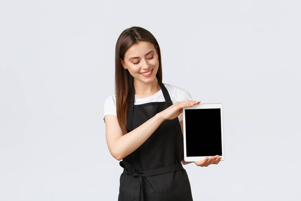 Grocery store employees, small business and coffee shops concept. Friendly cute saleswoman in black apron showing digital tablet screen and smiling at gadget display, showing advertisement - Photo, Image