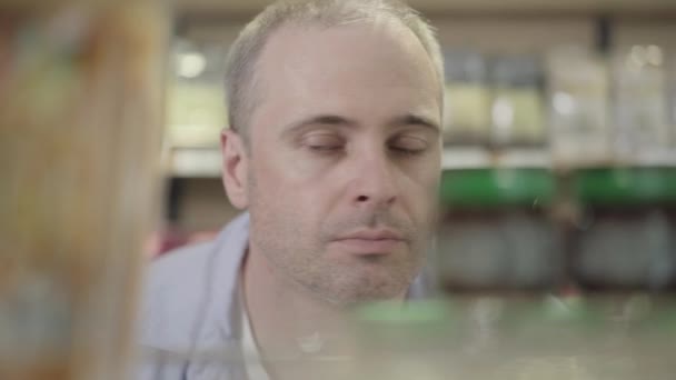Close-up face of focused confident man looking at canned food on shelf. Portrait of focused Caucasian buyer choosing products in grocery. Consumerism, healthy eating, lifestyle. - Video