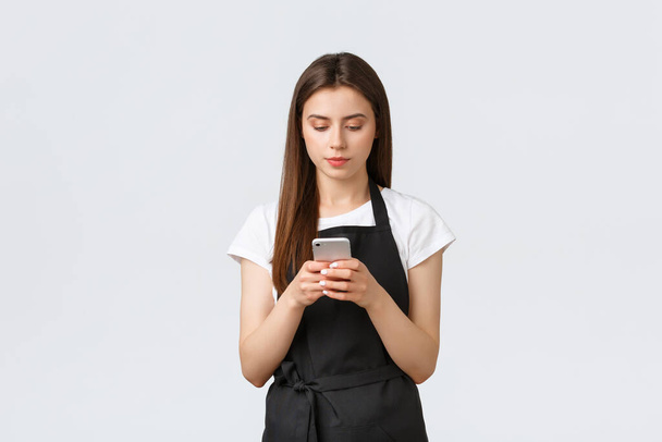 Grocery store employees, small business and coffee shops concept. Serious young female barista, waitress on break in black apron using smartphone, looking focused at screen - Foto, Bild