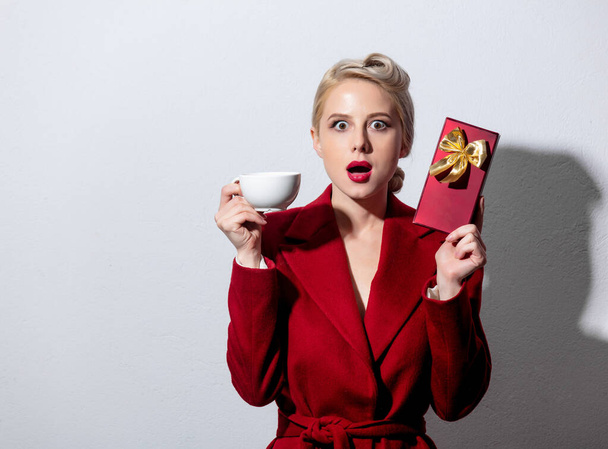 Szőke lány piros kabátban és vintage frizura csésze kávé és édességet doboz fehér háttér - Fotó, kép