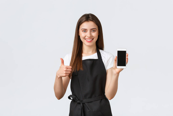 Grocery store employees, small business and coffee shops concept. Satisfied cute friendly-looking barista or waitress show thumbs-up and mobile phone sreen, recommend download application - Foto, immagini