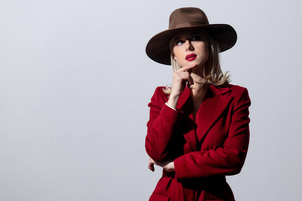 Szőke lány, piros kabátban és vintage kalapban, szürke háttérrel. Kép noir stílusban  - Fotó, kép