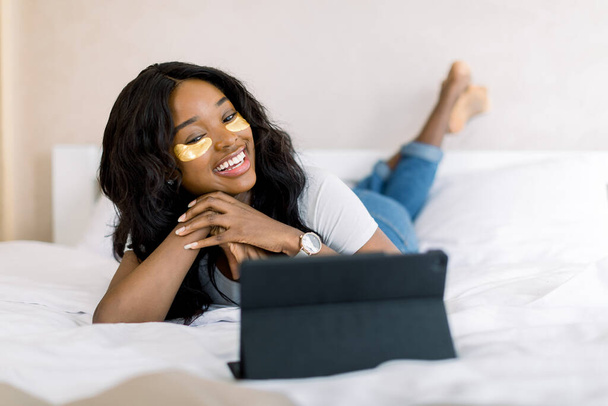 Relaks i zabiegi kosmetyczne w weekend. Przyjemna afrykańska uśmiechnięta kobieta leżąca na łóżku w przytulnym pokoju, ze złotymi opaskami na oczach i oglądająca film na tablecie iPad lub prowadząca rozmowę wideo - Zdjęcie, obraz