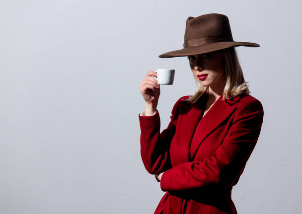 Blonde girl in red coat and vintage hat with cup of coffee on grey background. Image in noir style - Photo, Image