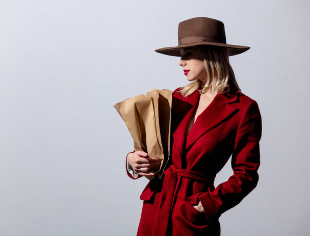 Blonde girl in red coat and vintage hat with paper supermarket bag on grey background. Image in noir style - Photo, Image