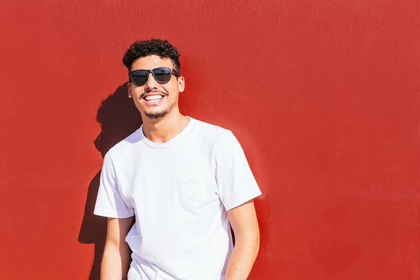 портрет молодого человека в солнечных очках, улыбающегося на красной стене
 - Фото, изображение
