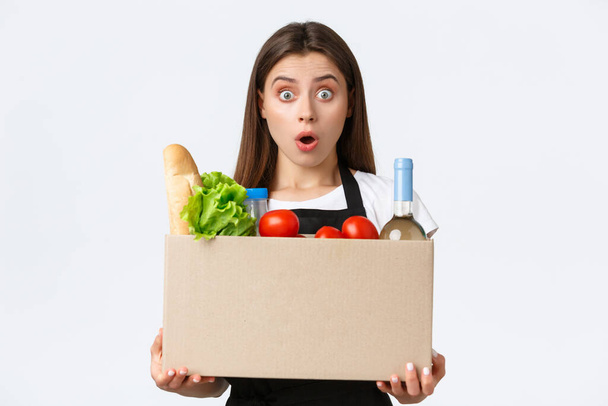 Employees, delivery and online orders, grocery stores concept. Shocked cashier or saleswoman holding box with food and groceries, looking amazed with dropped jaw, provide home-deliver - Photo, Image