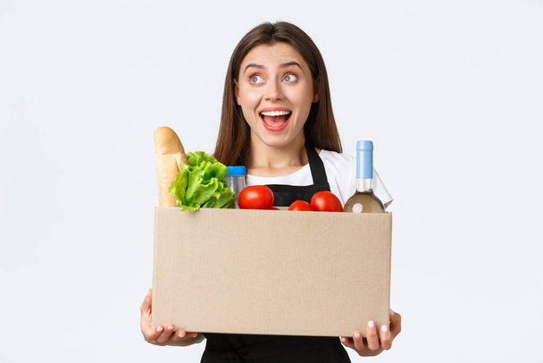 Employees, delivery and online orders, grocery stores concept. Smiling cheerful female courier, saleswoman packed online order in box, holding package with groceries and turn left pleased - Photo, Image