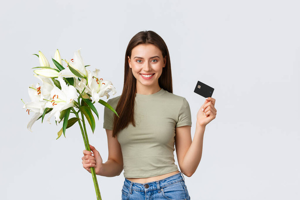 Vrolijke tedere jonge vrouw bestelde boeket bloemen levering met creditcard, glimlachend blij en met mooie witte lelies. Meisje betalen voor aankoop in internet, staande witte achtergrond - Foto, afbeelding