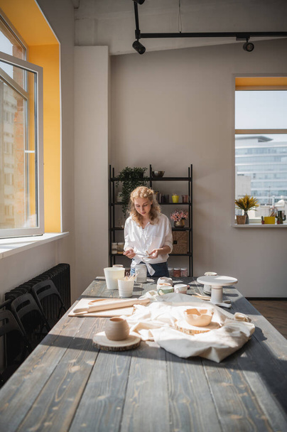 Egy fiatal európai gyönyörű női fazekas egy kreatív folyamat, hogy kerámia asztalnál a műhelyben. Haj repked a szélben a napsütéses napon a fazekas stúdióban. - Fotó, kép