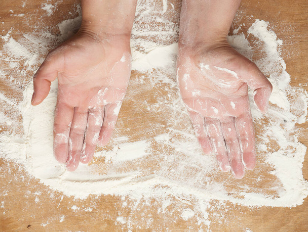 two hands and white wheat flour scattered on a brown wooden background, product is spread over the surface, kitchen background, top view - Fotografie, Obrázek