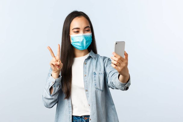 Koncepcja społecznego dystansu, pandemii covid-19 i ludzkich emocji. Przyjazny wesoły azjatycki kobieta w medycznej masce robi selfie dla mediów społecznościowych, zrobić znak pokoju w aparacie smartfona - Zdjęcie, obraz