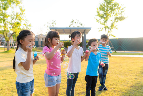 Large group of happy Asian smiling kindergarten kids friends playing blowing bubbles together in the park on the green grass on sunny summer day. - Photo, image