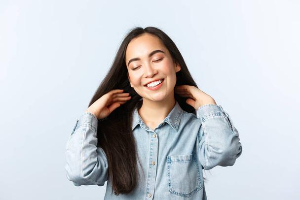 Lifestyle, people emotions and beauty concept. Carefree attractive asian girl taking care of long dark hair, show-off beautiful hairstyle, done new haircut, close eyes and smiling satisfied - Valokuva, kuva