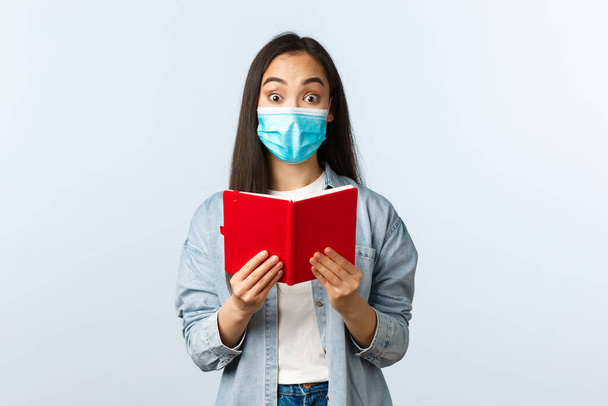 Covid-19 pandemic, education during coronavirus, back to school concept. Cheerful friendly smiling asian woman in medical mask prepare for college exam, studying notes in notebook - Photo, image