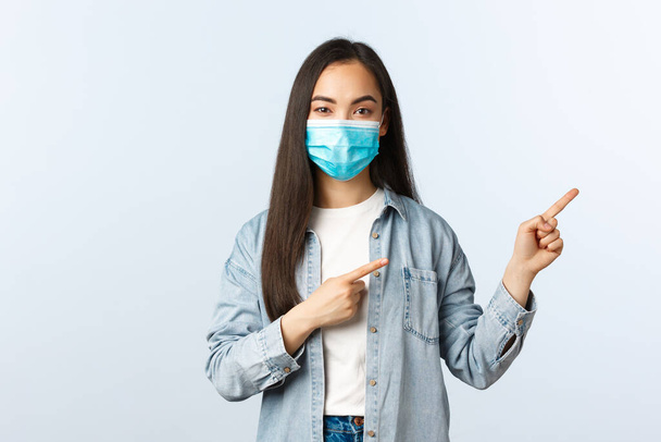 Social distancing lifestyle, covid-19 pandemic everyday life and leisure concept. Confident attractive female asian student in medical mask provide information, pointing fingers right - 写真・画像