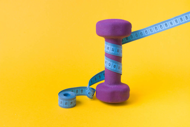 1.5 kilogram purple dumbbell and blue measuring tape on a yellow background, a dumbbell wrapped in a folded tape copy space, weight loss concept - Φωτογραφία, εικόνα
