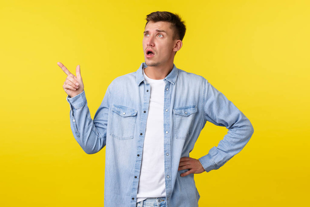 Confused and surprised startled handsome adult man asking question about product, looking and pointing finger upper left corner, consulting shop clerk, standing yellow background - Fotó, kép