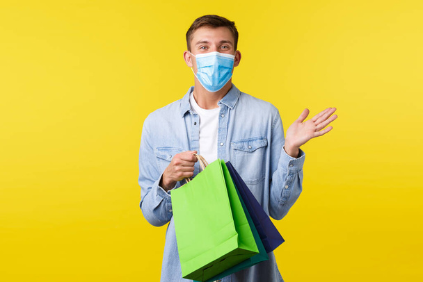 Concept of covid-19 pandemic outbreak, shopping and lifestyle during coronavirus. Handsome happy young man in medical mask enjoying buying things in store, holding bags with presents - Foto, imagen