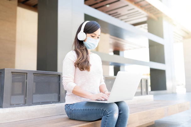 femme dans le masque facial à l'aide d'un ordinateur portable assis sur le siège dans la ville pendant l'épidémie de coronavirus - Photo, image