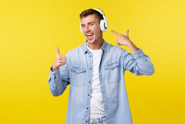 Lifestyle, summer holidays, technology concept. Happy handsome man, student in wireless headphones pointing at earphones and show thumbs-up as satisfied with good music, awesome beats - Foto, Imagem
