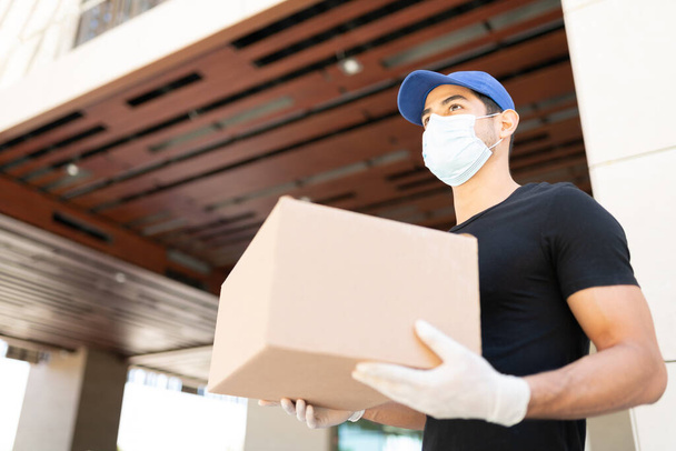 Hispanic young delivery man wearing face mask while carrying parcel box during coronavirus outbreak. Focus on man - Photo, Image