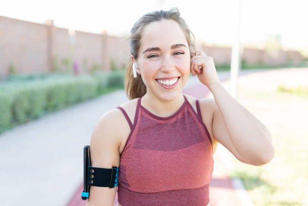 Sonriente joven atractiva escuchando música a través de auriculares inalámbricos mientras hace ejercicio en el parque
 - Foto, Imagen