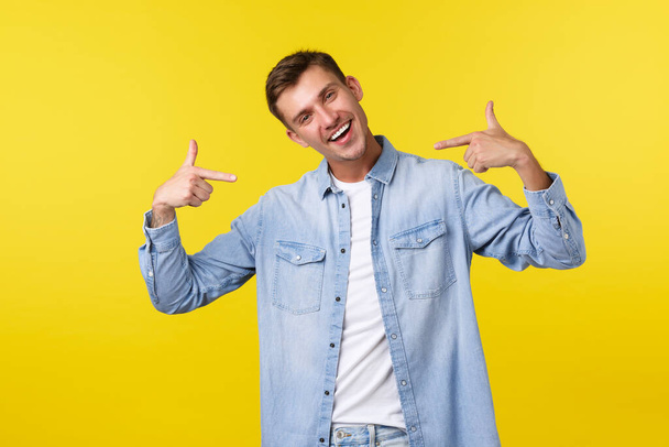 Portrait of happy handsome blond male student pointing at himself and smiling joyfully. Guy show-off, bragging over personal achievement, being excited as got job or promotion, yellow background - Photo, Image