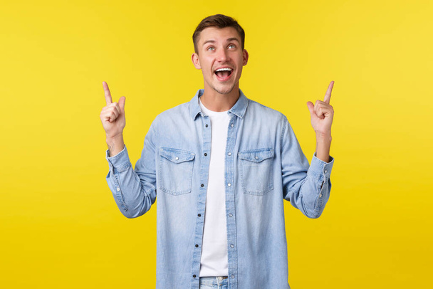 Enthusiatic happy handsome guy laughing and smiling broadly, looking and pointing fingers up, reading funny banner, receive special summer discounts, ready for shopping over yellow background - Photo, Image