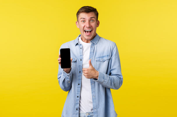 Szabadidő, technológia és alkalmazás reklám koncepció. Izgalmas boldog és elégedett szőke srác mutatja hüvelykujj-up, mint ajánlani szuper cool új app, mutatja online kedvezmények mobil képernyőn - Fotó, kép