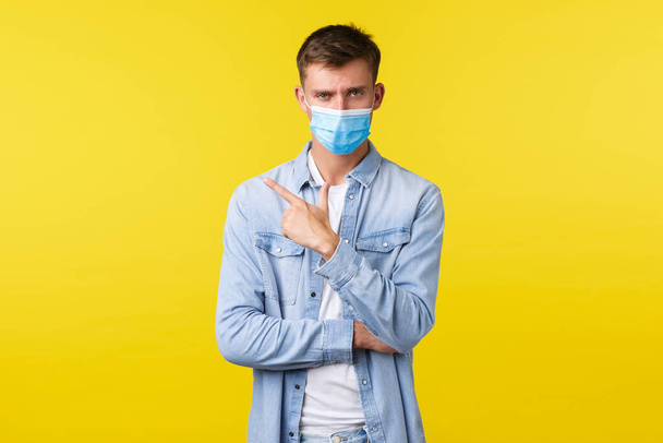 Concept of covid-19 pandemic outbreak, lifestyle during coronavirus social distancing. Serious looking concerned man in medical mask, pointing finger upper left corner, yellow background - Photo, Image