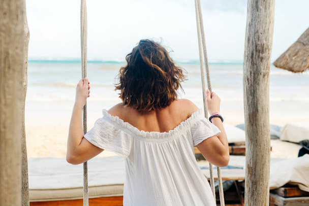Mädchen in weißem Kleid schwingt auf einer Schaukel seinen Rücken am Strand. - Foto, Bild
