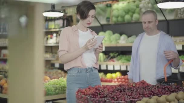 Positive Caucasian man and woman touching hands accidentally as taking strawberry from supermarket shelf. Portrait of laughing male and female customer buying food in grocery. Lifestyle, flirt. - Materiał filmowy, wideo