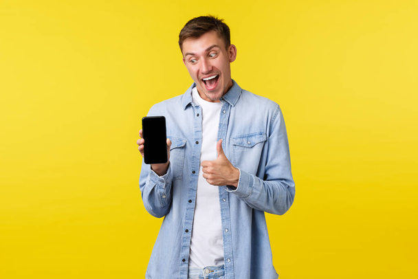 Leisure, technology and application advertisement concept. Happy excited blond guy show thumbs-up as looking and pointing at mobile phone display, checking out new cool promo or app - Foto, Imagem