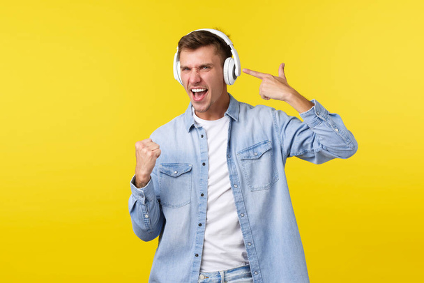 Lifestyle, summer holidays, technology concept. Upbeat handsome man in casual clothes listening music in headphones, pointing at earphone as enjoying nice beats and awesome song, yellow background - Photo, Image